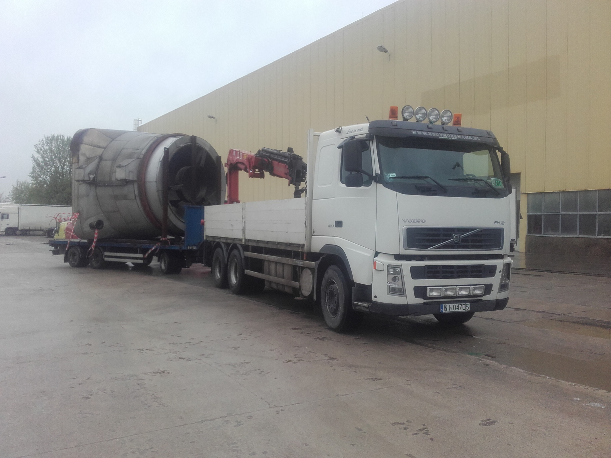 Biała ciężarówka Volvo z czerwoną mini-żurawiem na pace, transportująca na niskopodwoziowej przyczepie duży, metalowy, cylindryczny zbiornik, na tle hali przemysłowej w pochmurny dzień.