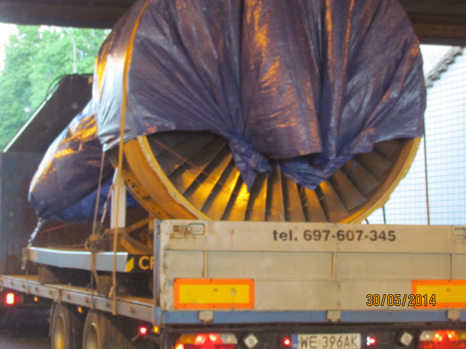 Duża turbina, częściowo przykryta niebieską plandeką, transportowana na platformie ciężarówki. Widoczny numer telefonu na burcie i data 30/05/2014.