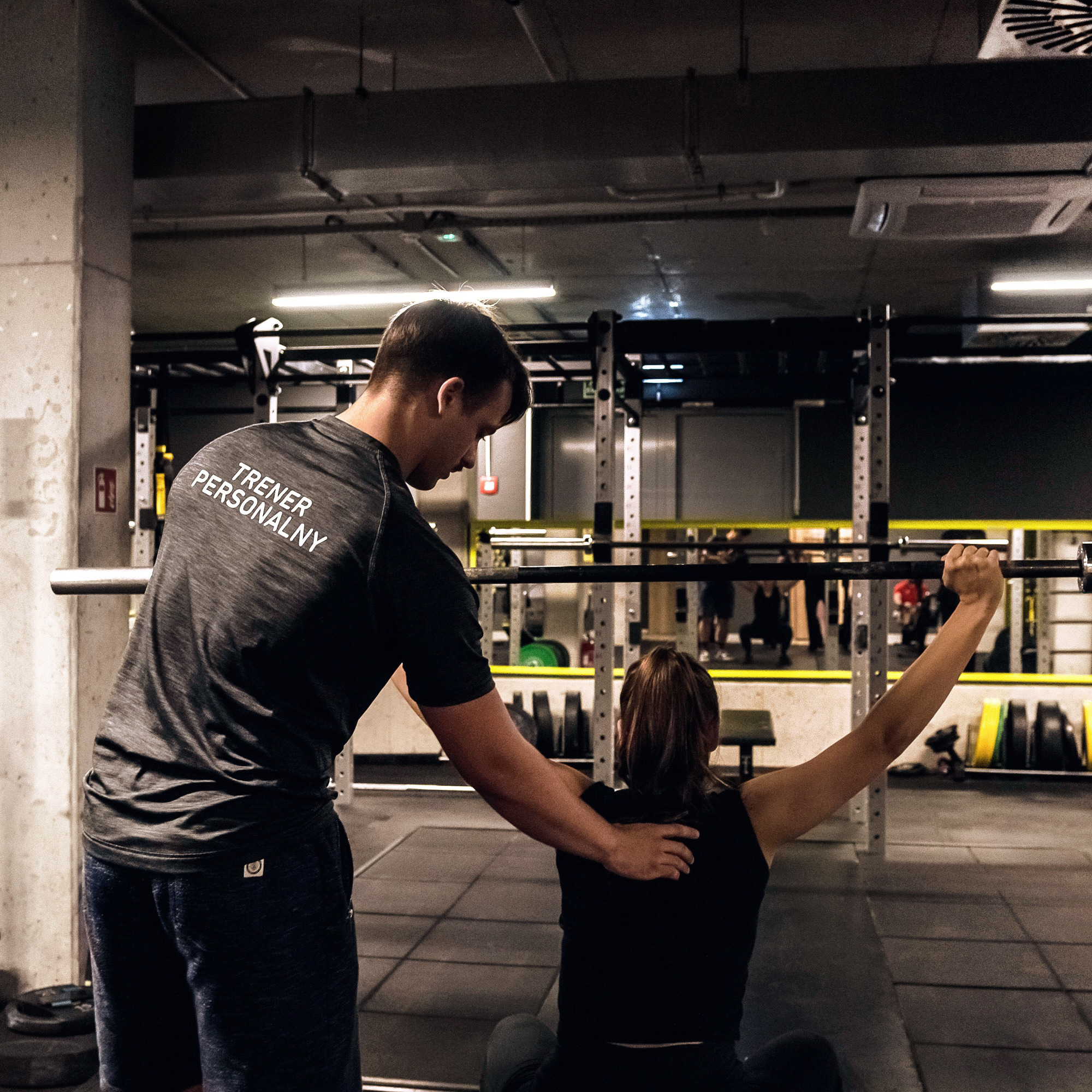 Kobieta ćwiczy podciąganie na maszynie pod okiem mężczyzny z napisem na plecach koszulki. Sprzęt do ćwiczeń siłowych w tle.