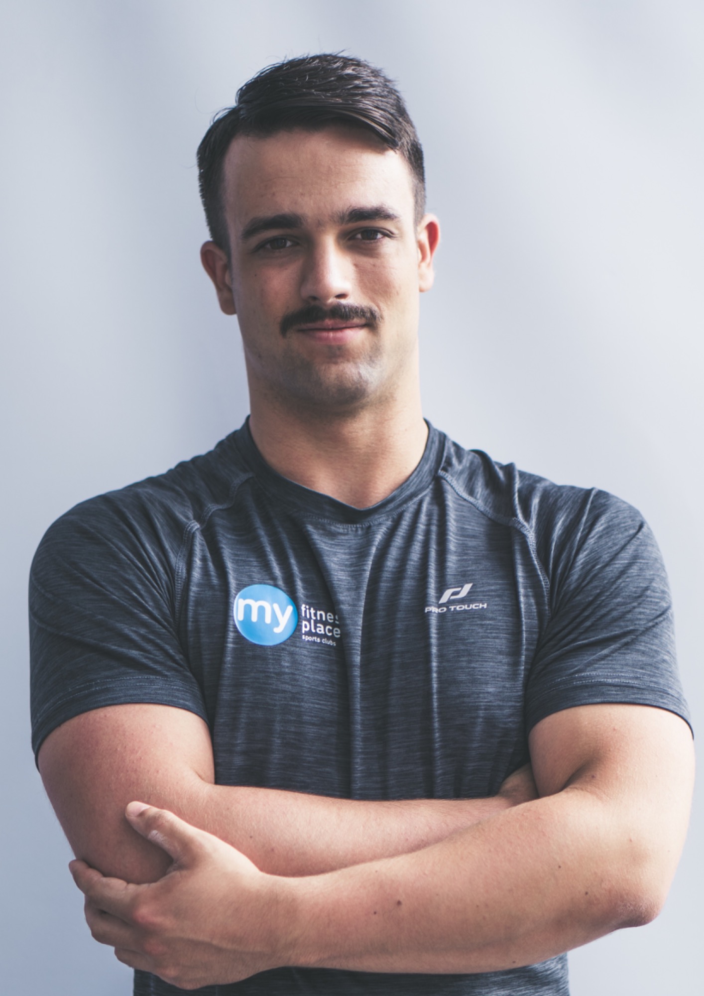 Portret mężczyzny z wąsami, ubranego w sportową koszulkę z logo 'My Fitness Place', pozującego z założonymi rękami na tle szarej ściany.