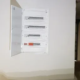 Otwarta szafka rozdzielcza z zamontowanymi bezpiecznikami na ścianie w trakcie prac wykończeniowych, widoczne ślady po malowaniu.