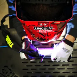 Widok z góry na osobę w masce spawalniczej Lincoln Electric, pracującą nad metalową konstrukcją z widocznym palnikiem TIG i intensywnym światłem łuku.