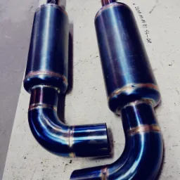 Dwa identyczne, niebiesko-złote tłumiki spalin z widocznymi spawami na stole warsztatowym, z notatką na stole.