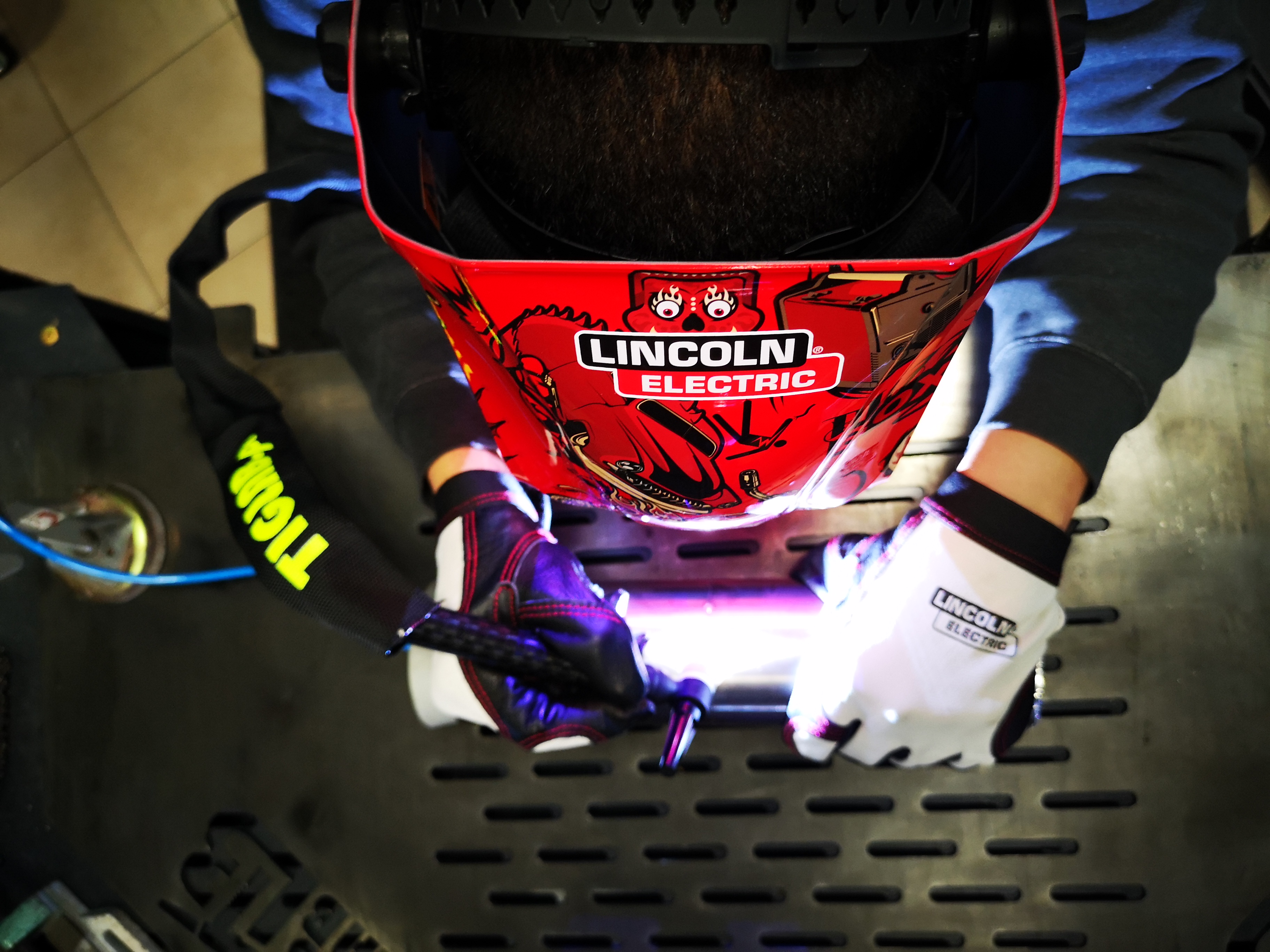 Widok z góry na osobę w masce spawalniczej Lincoln Electric, pracującą nad metalową konstrukcją z widocznym palnikiem TIG i intensywnym światłem łuku.