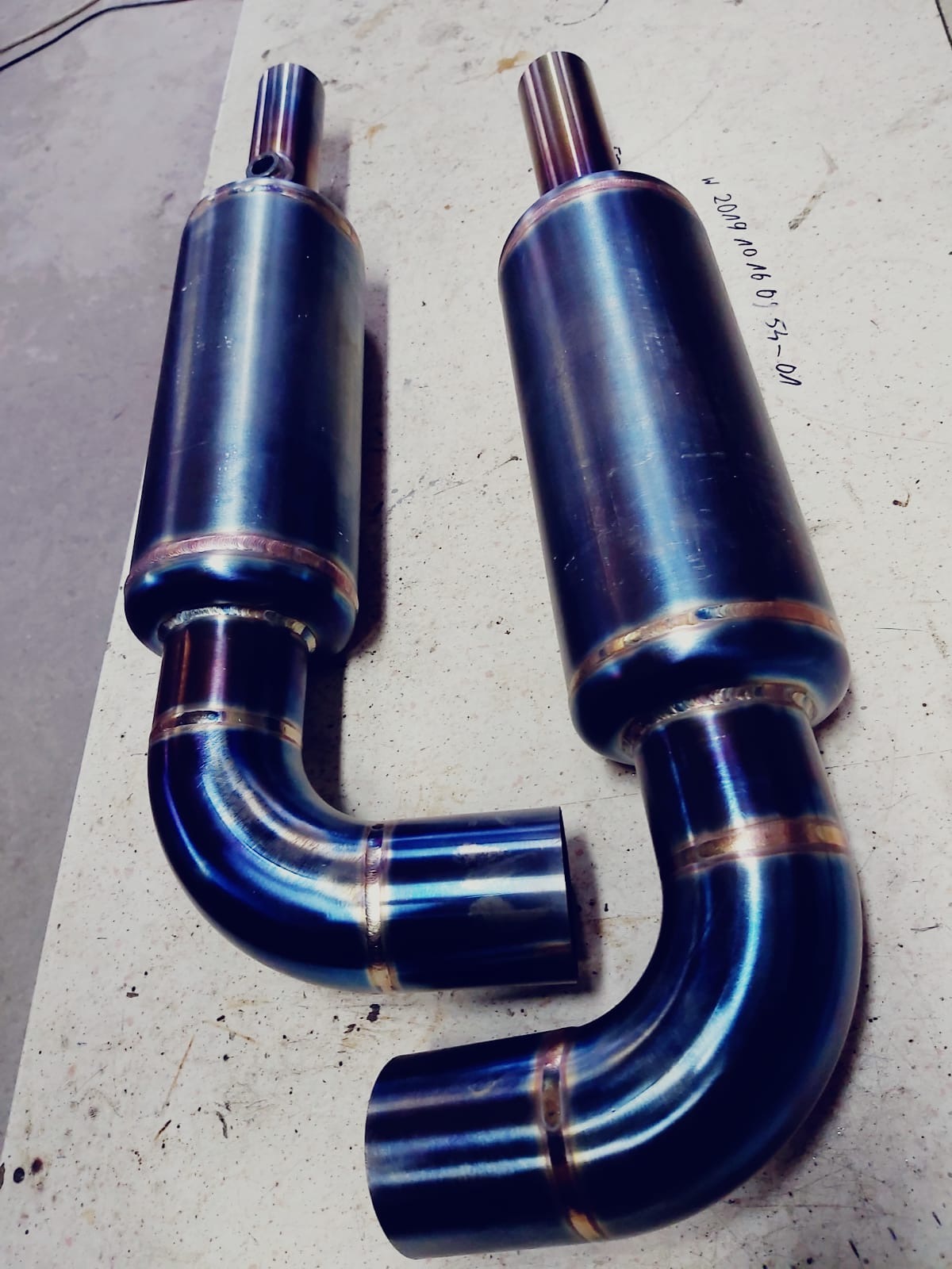 Dwa identyczne, niebiesko-złote tłumiki spalin z widocznymi spawami na stole warsztatowym, z notatką na stole.