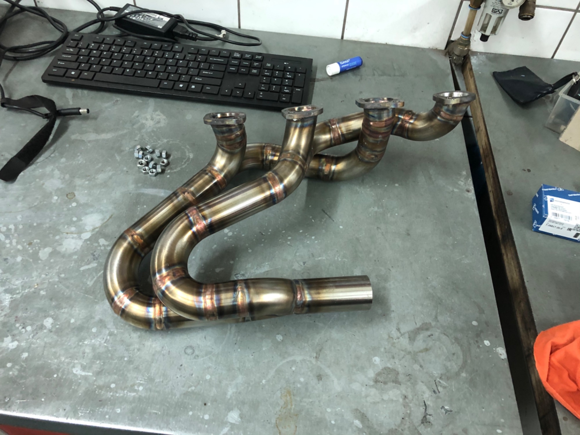 Kolektor wydechowy ze stali nierdzewnej, spawany metodą TIG, leżący na warsztatowym stole obok klawiatury komputerowej i drobnych elementów montażowych.