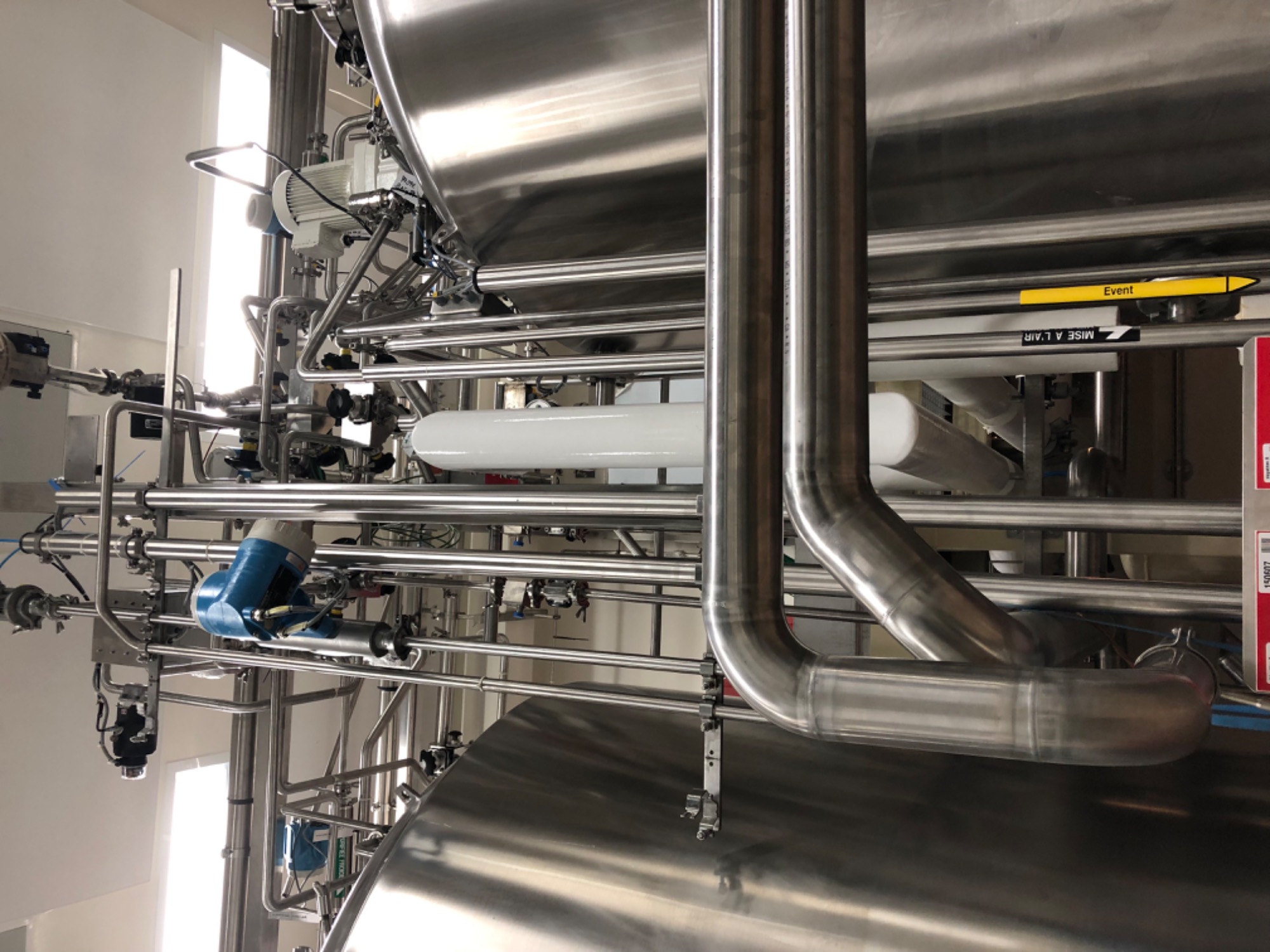 Kompleksowa instalacja przemysłowa z rur ze stali nierdzewnej, zbiorników i zaworów, widoczne żółte i czarne etykiety informacyjne.