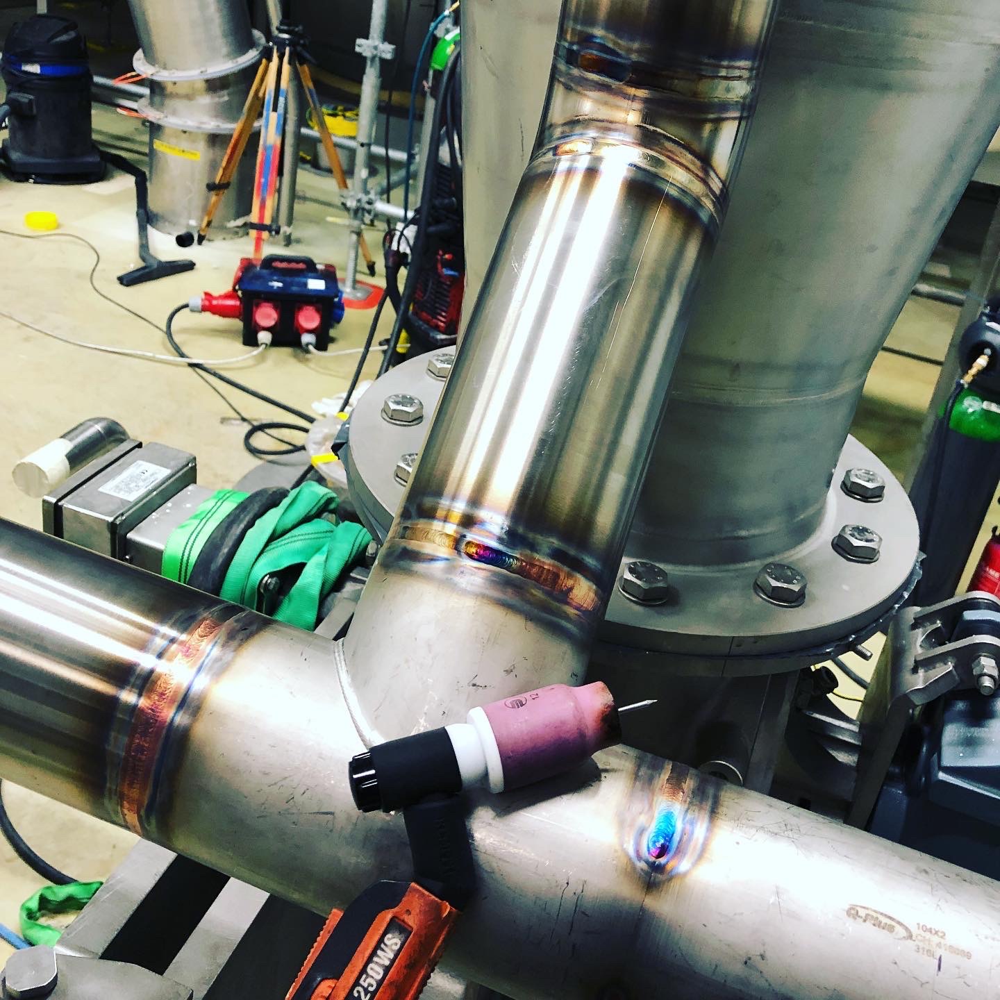 Precyzyjne łączenie rur ze stali nierdzewnej metodą TIG, widoczne tęczowe przebarwienia spoiny oraz uchwyt spawalniczy z różową dyszą ceramiczną.