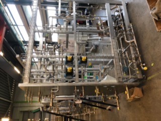 Skomplikowana konstrukcja ze stali nierdzewnej, składająca się z rur, zaworów i zbiorników, umieszczona na metalowej ramie. Widoczne elementy elektryczne w żółtych obudowach. Zdjęcie wykonane...