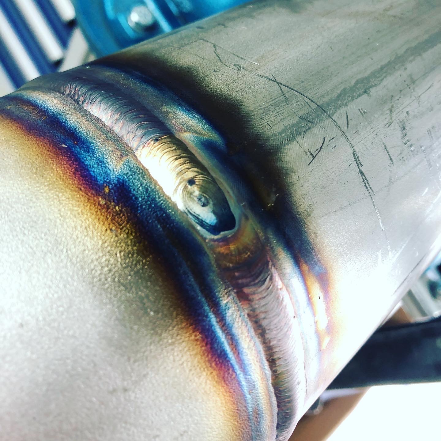 Szczegółowe ujęcie spoiny na metalowej rurze, widoczne tęczowe przebarwienia termiczne i delikatne rysy na powierzchni.