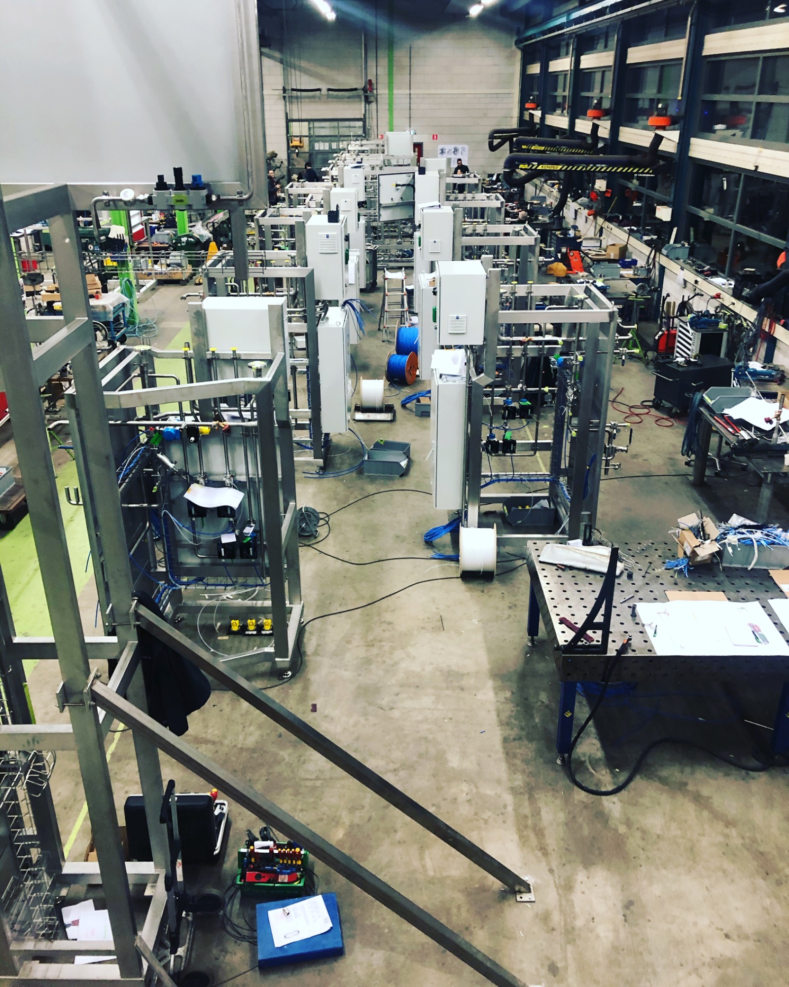 Szeroki kadr na halę produkcyjną z licznymi, metalowymi konstrukcjami w trakcie montażu, widoczne skrzynki elektryczne, przewody, narzędzia i stanowiska pracy, z góry