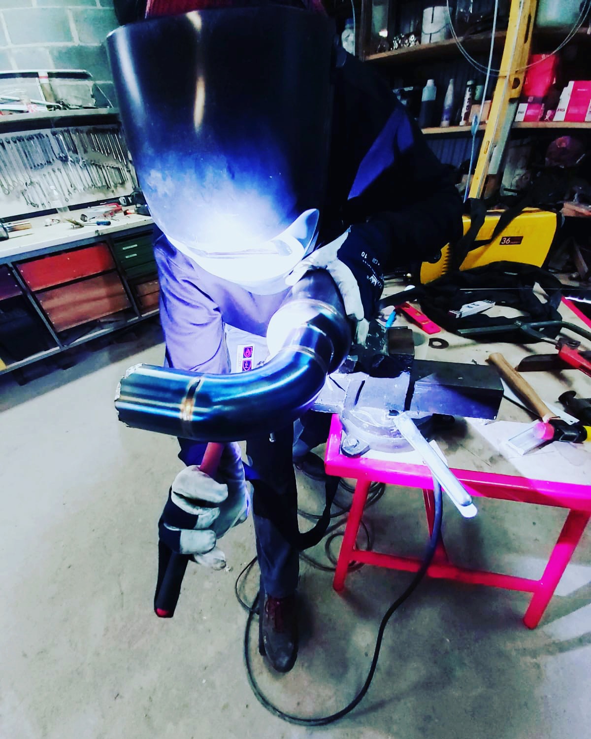 Praca w warsztacie: spawanie rury ze stali nierdzewnej w osłonie argonu, widoczna jasna iskra i niebieska poświata na masce ochronnej.