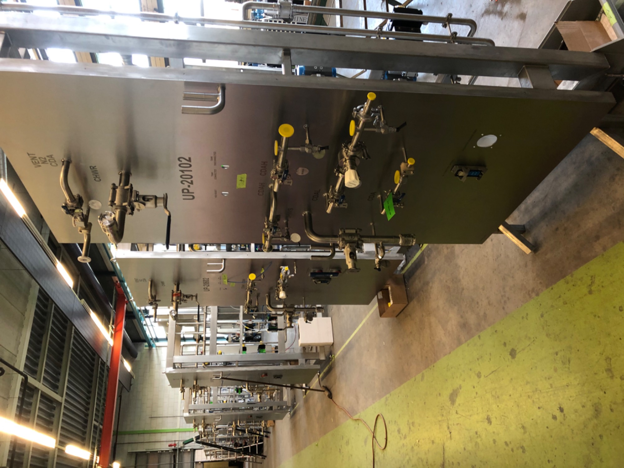 Skomplikowany system rur i zaworów ze stali nierdzewnej zamontowany na panelach, z widocznymi oznaczeniami 'UP-20102' i 'UP-20602', zainstalowany w hali produkcyjnej z widoczną instalacją...