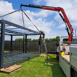 Montaż stalowej konstrukcji szkieletowej przy użyciu dźwigu samochodowego Fassi F50 na tle zielonej trawy i błękitnego nieba.