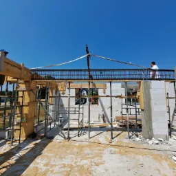 Wznoszenie ścian domu z betonu komórkowego, widoczne szalunki i zbrojenia, dwóch pracowników na budowie w słoneczny dzień.