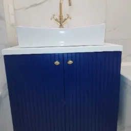 Łazienkowa szafka pod umywalkę w odcieniu głębokiego granatu z pionowymi żłobieniami, biały blat i prostokątna umywalka nablatowa, złota bateria z dwoma kurkami.