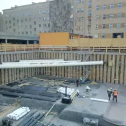 Głęboki wykop pod budowę otoczony ścianą z pali, na dnie składowane pręty zbrojeniowe i białe rury, w tle budynki mieszkalne i pracownicy w odblaskowych kamizelkach.