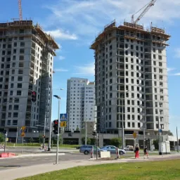 Dwa wysokie budynki w stanie surowym otwartym, z żurawiami na szczycie, widoczne z poziomu ulicy z przejeżdżającym autobusem i przechodniami.