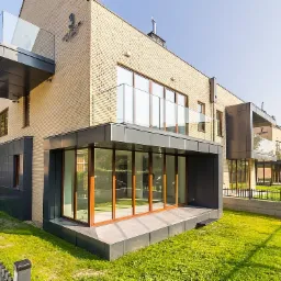 Nowoczesny budynek wielorodzinny z jasnej cegły z przeszklonymi balkonami i tarasami, otoczony zielonym trawnikiem i ogrodzeniem.