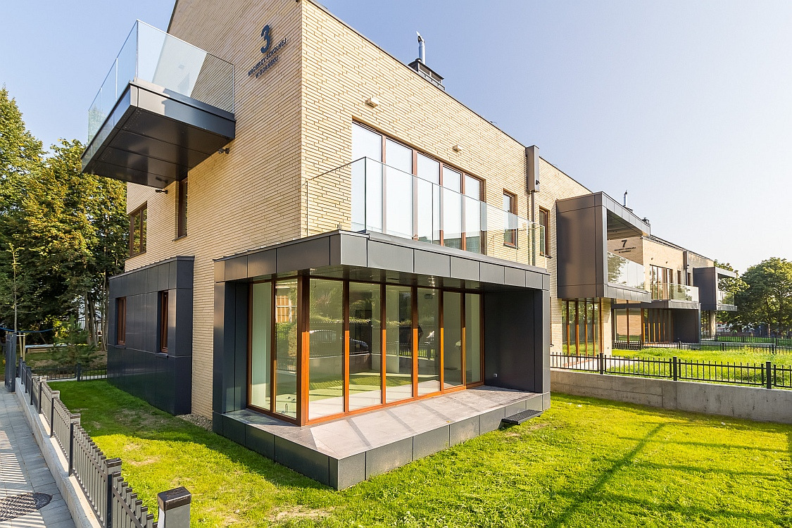 Nowoczesny budynek wielorodzinny z jasnej cegły z przeszklonymi balkonami i tarasami, otoczony zielonym trawnikiem i ogrodzeniem.