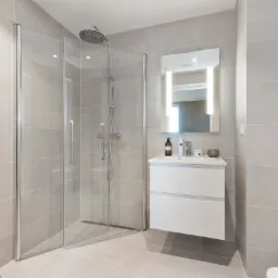 Nowoczesna łazienka z szarą płytką, kabiną prysznicową typu walk-in, umywalką z szafką i podświetlanym lustrem, uzupełniona o pomarańczowy ręcznik na wieszaku.