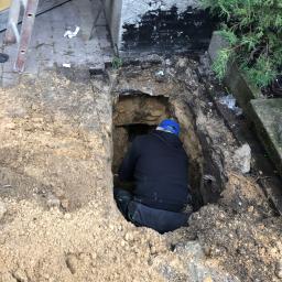 Tomi Roboty Ziemne - Pracownik w niebieskiej czapce w głębokim wykopie ziemnym obok budynku z drabiną opartą o ścianę, prawdopodobnie podczas prac związanych z instalacjami podziemnymi.