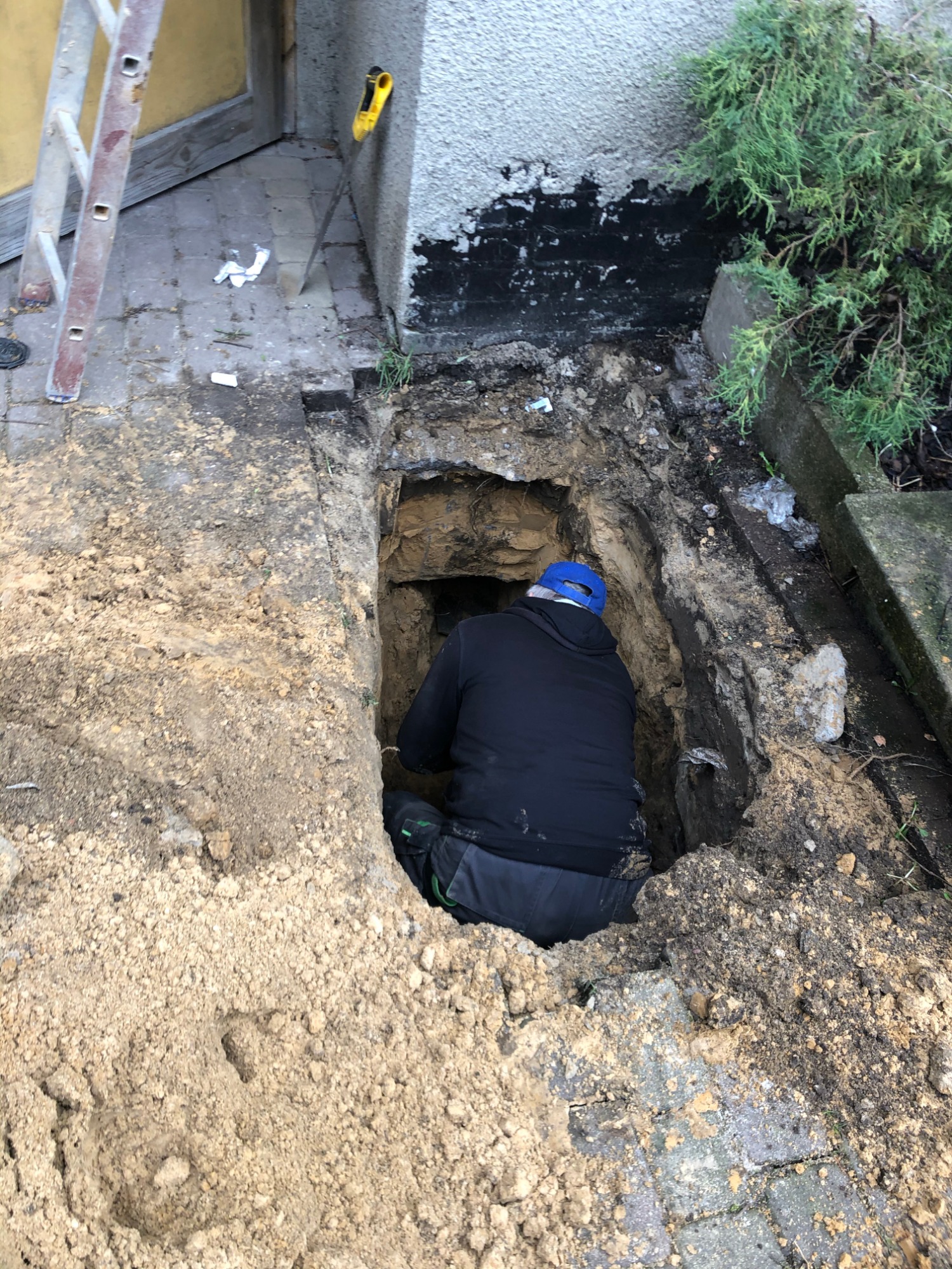 Pracownik w niebieskiej czapce w głębokim wykopie ziemnym obok budynku z drabiną opartą o ścianę, prawdopodobnie podczas prac związanych z instalacjami podziemnymi.