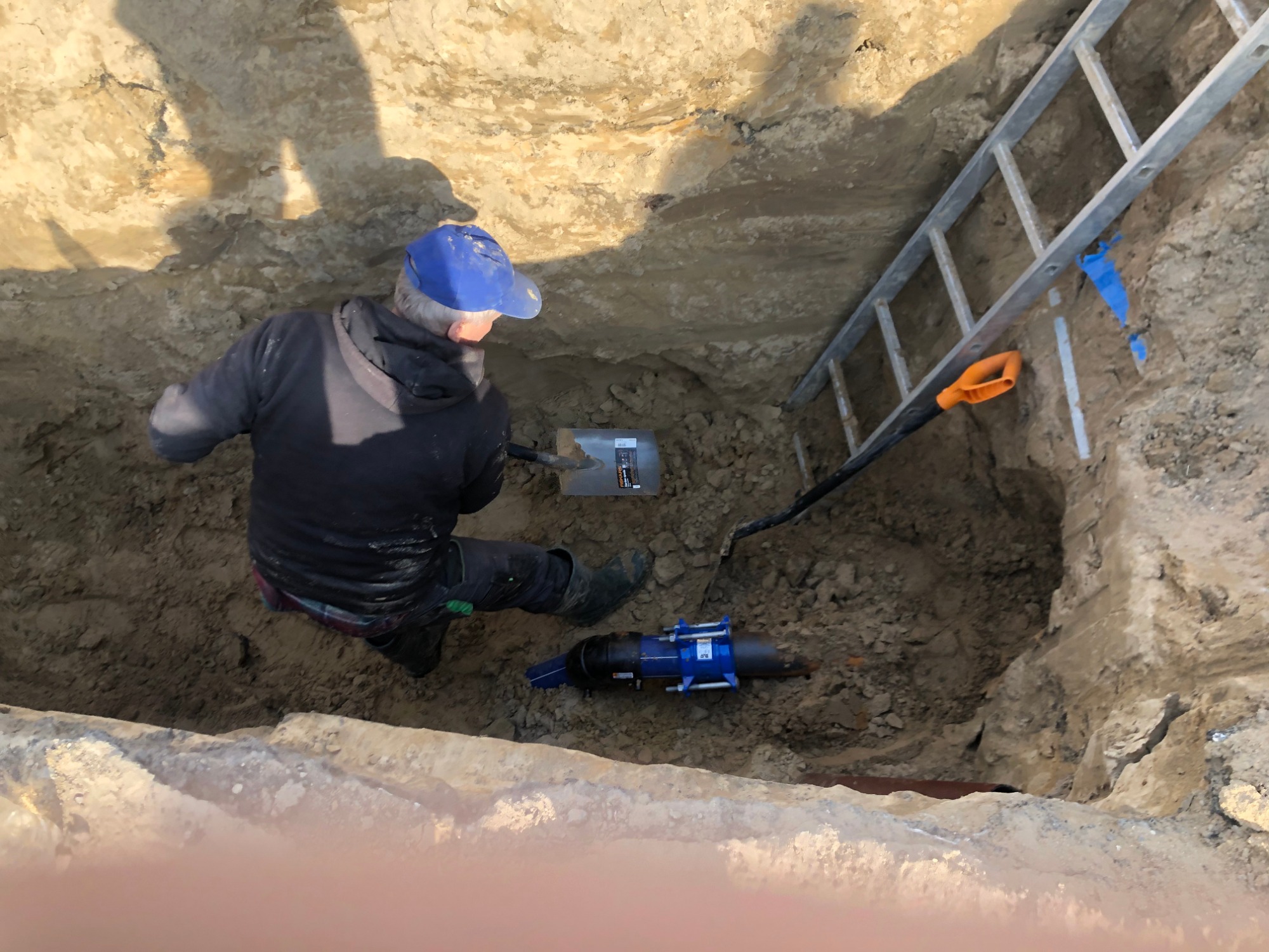 Pracownik w wykopie, w niebieskiej czapce, kopie łopatą obok niebieskiego narzędzia do łączenia rur, z metalową drabinką opartą o krawędź wykopu.