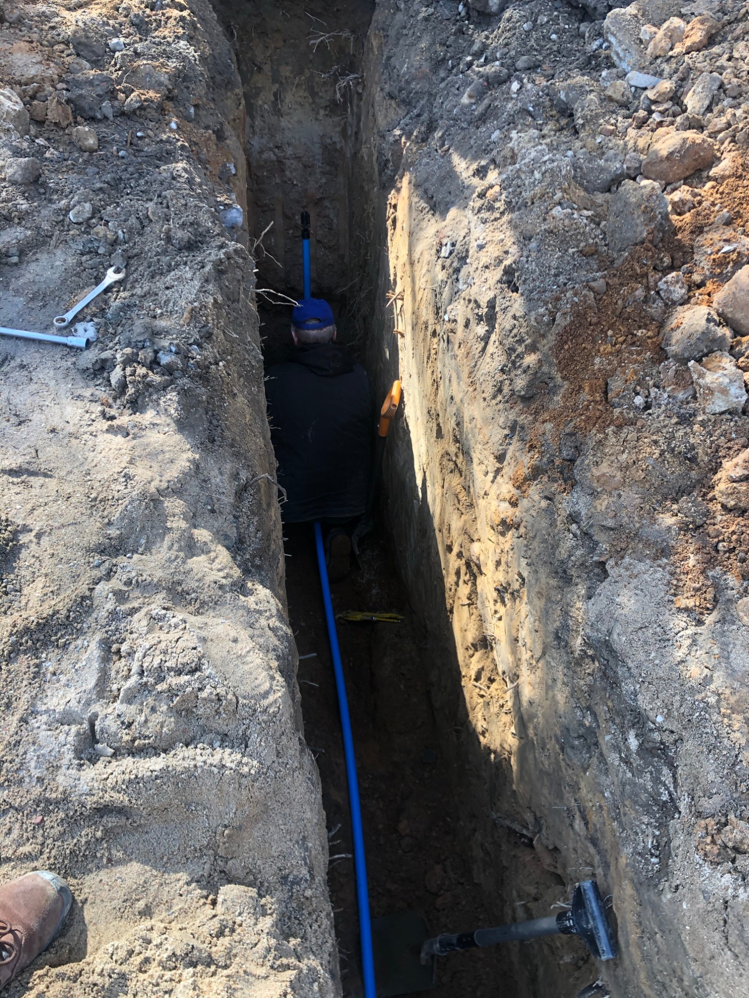 Wykop z ułożoną niebieską rurą, widoczny pracownik w wykopie, narzędzia i ziemia.