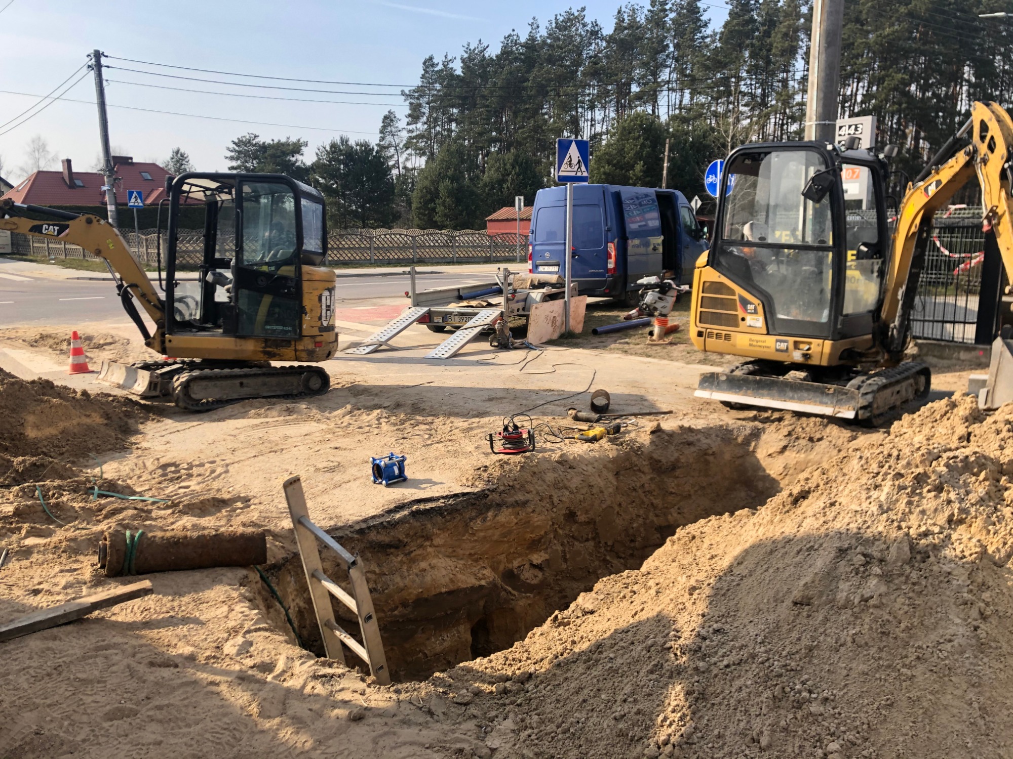 Dwa mini-koparki CAT po obu stronach wykopu w piaszczystej ziemi, widoczna rura kanalizacyjna, drabina, niebieski van i sprzęt na przyczepie w tle.