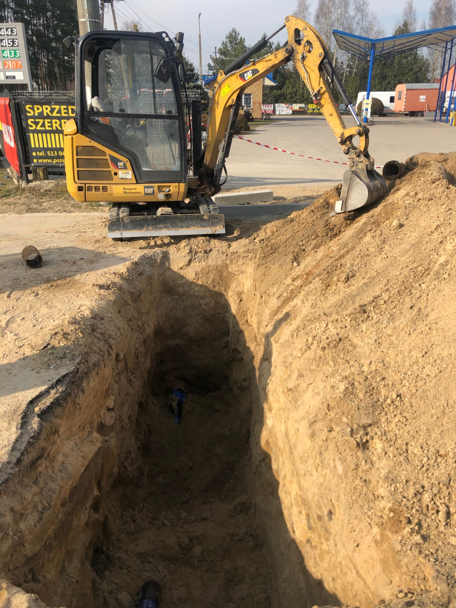 Żółta mini koparka CAT przy wykopie w ziemi, wewnątrz widoczne niebieskie rury, na pierwszym planie fragment rury, w tle budynek stacji paliw.