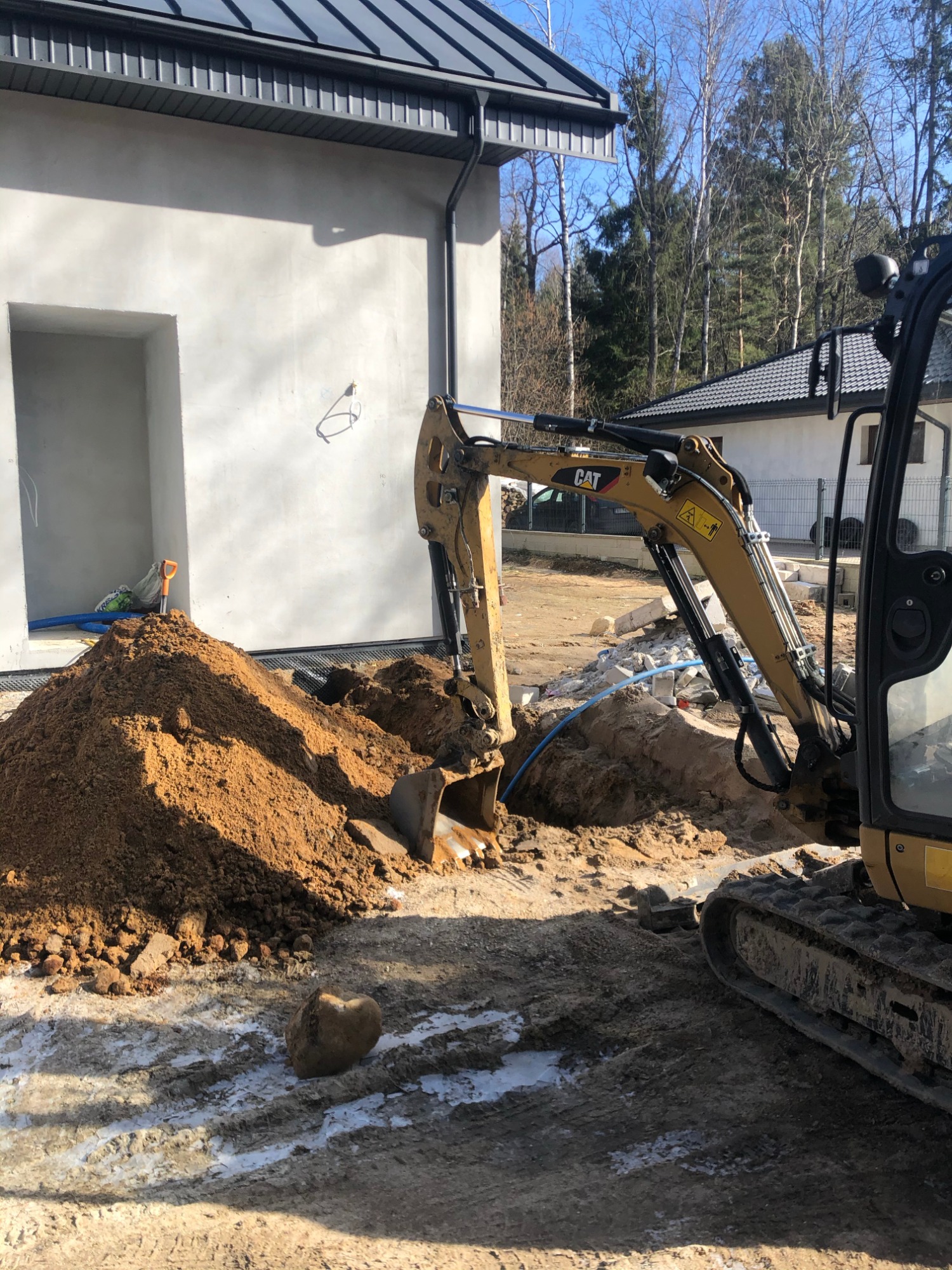 Żółta koparka CAT wykonuje wykop pod przyłącze do nowo budowanego domu jednorodzinnego, widoczny fragment rury i sterta ziemi z łopatą.