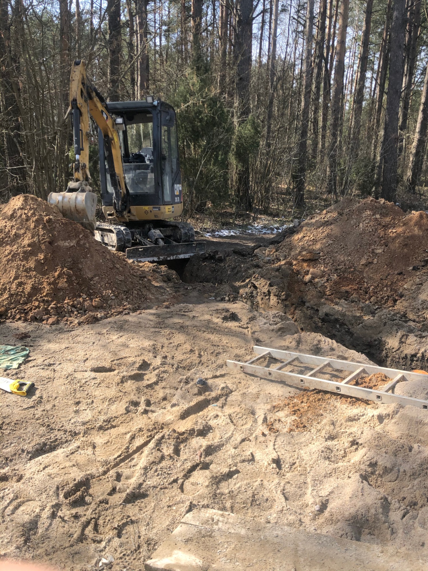 Żółta minikoparka CAT 301.7D w trakcie wykonywania wykopu pod przyłącze do domu jednorodzinnego, widoczna drabina aluminiowa i poziomica na piasku.