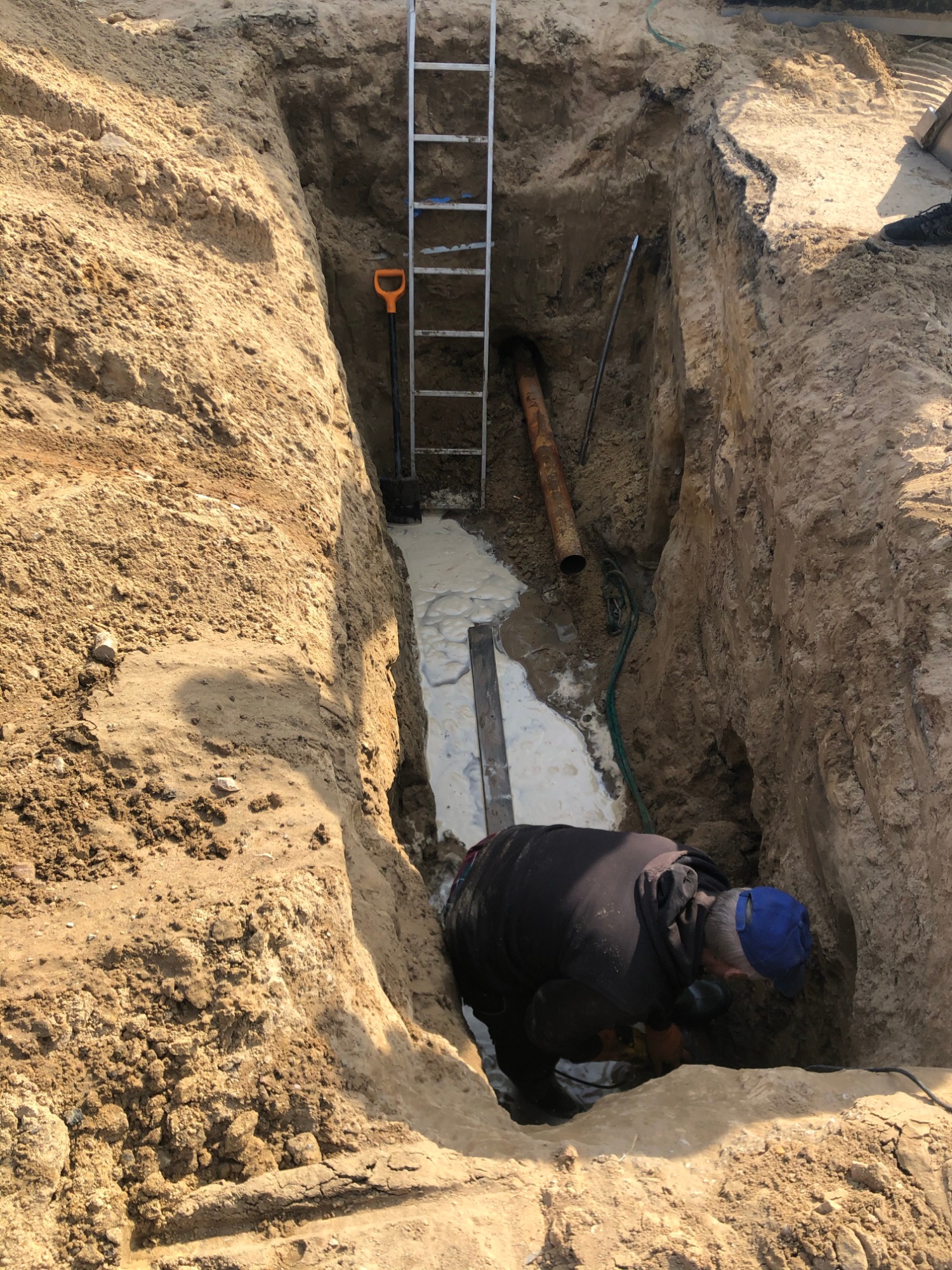 Pracownik w wykopie, przyłącze rury kanalizacyjnej, widoczna drabina, szpadel i zalewająca wykop woda.