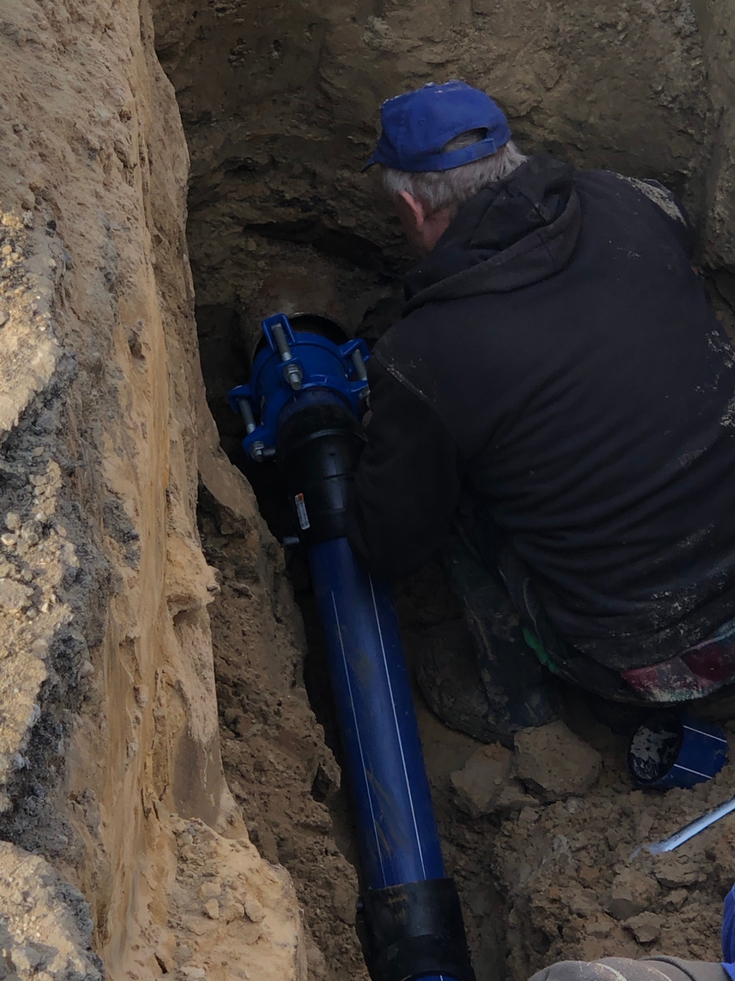 Pracownik w wykopie łączy niebieską rurę z elementem przyłączeniowym, widoczne ślady ziemi na ubraniu.