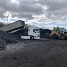 Ciężarówka wyładowująca czarny materiał sypki na placu, obok pracuje ładowarka kołowa, widoczne betonowe słupki i pochmurne niebo.