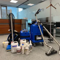 Profesjonalny sprzęt do czyszczenia dywanów w biurze, obejmujący odkurzacz piorący, szczotki, środki czyszczące marki Prochem i Santoemma, z widocznym w tle biurowym wnętrzem z przeszklonymi...