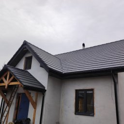 PPHU Czesław Gołębiewski - Nowoczesny dom jednorodzinny w trakcie wykończenia, z szarą dachówką ceramiczną i drewnianą konstrukcją zadaszenia wejścia, widoczne drabiny i materiały budowlane.