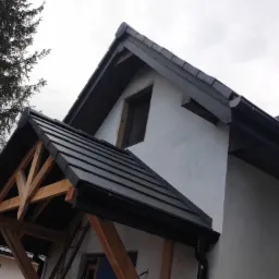 Nowe, ciemne pokrycie dachowe z drewnianą konstrukcją wejścia, widoczne rynny i fragment elewacji domu.