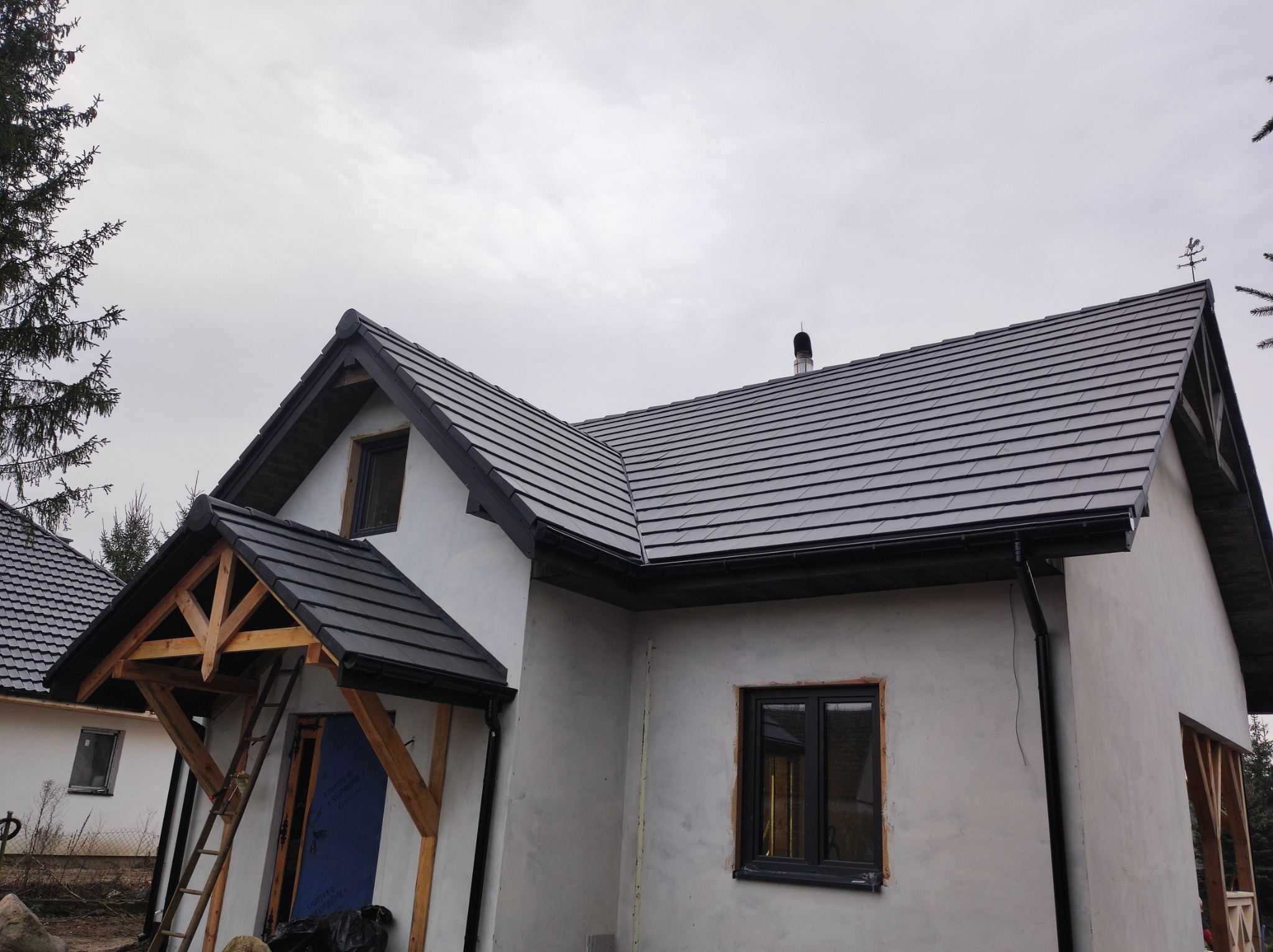 Nowoczesny dom jednorodzinny w trakcie wykończenia, z szarą dachówką ceramiczną i drewnianą konstrukcją zadaszenia wejścia, widoczne drabiny i materiały budowlane.