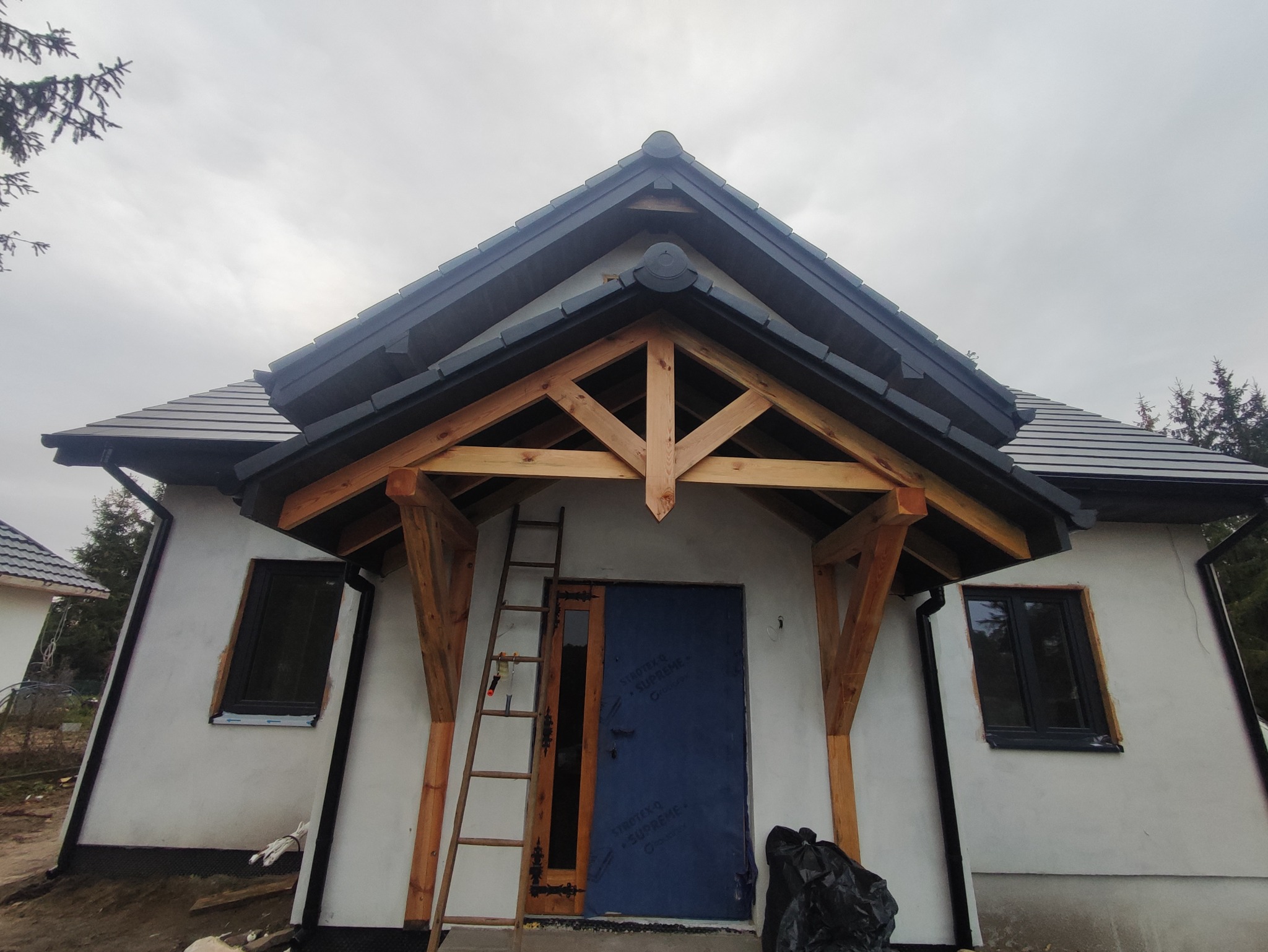 Nowy dom jednorodzinny w trakcie budowy, widoczny fragment elewacji z drewnianą konstrukcją wejścia, czarną stolarką okienną i pokryciem dachu z dachówki ceramicznej. Pod ścianą oparta drabina...