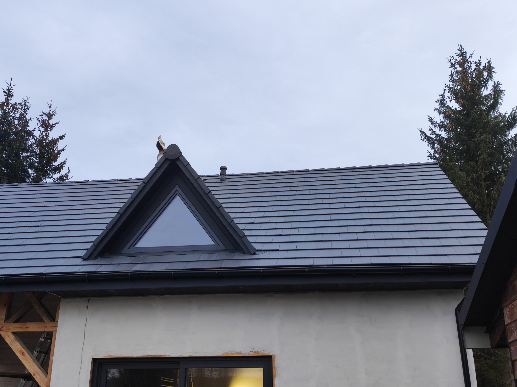 Szary dach z dachówki ceramicznej z trójkątnym oknem dachowym, widoczne elementy konstrukcji drewnianej domu w trakcie budowy.