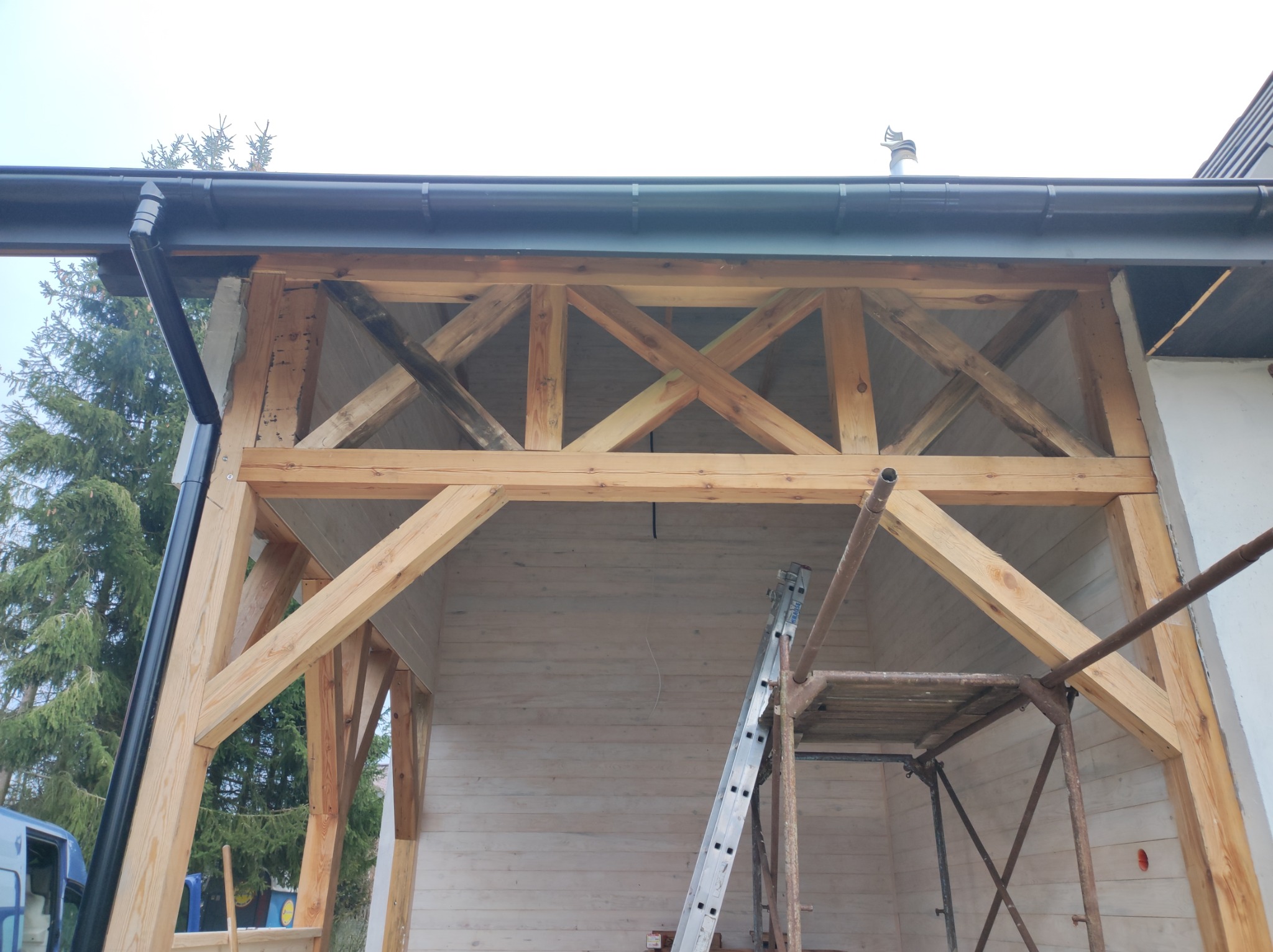 Konstrukcja drewniana z ukośnymi belkami podtrzymującymi dach, widoczne rusztowanie i drabina oparta o ścianę z desek, na zewnątrz widoczny fragment rynny i drzewo.