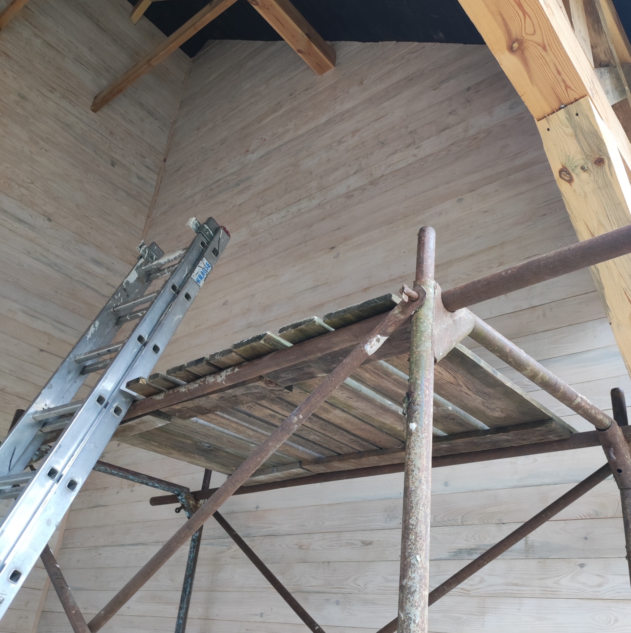 Wnętrze budynku w trakcie prac wykończeniowych, widoczne drewniane deski na ścianach, rusztowanie i drabina oparta o ścianę.