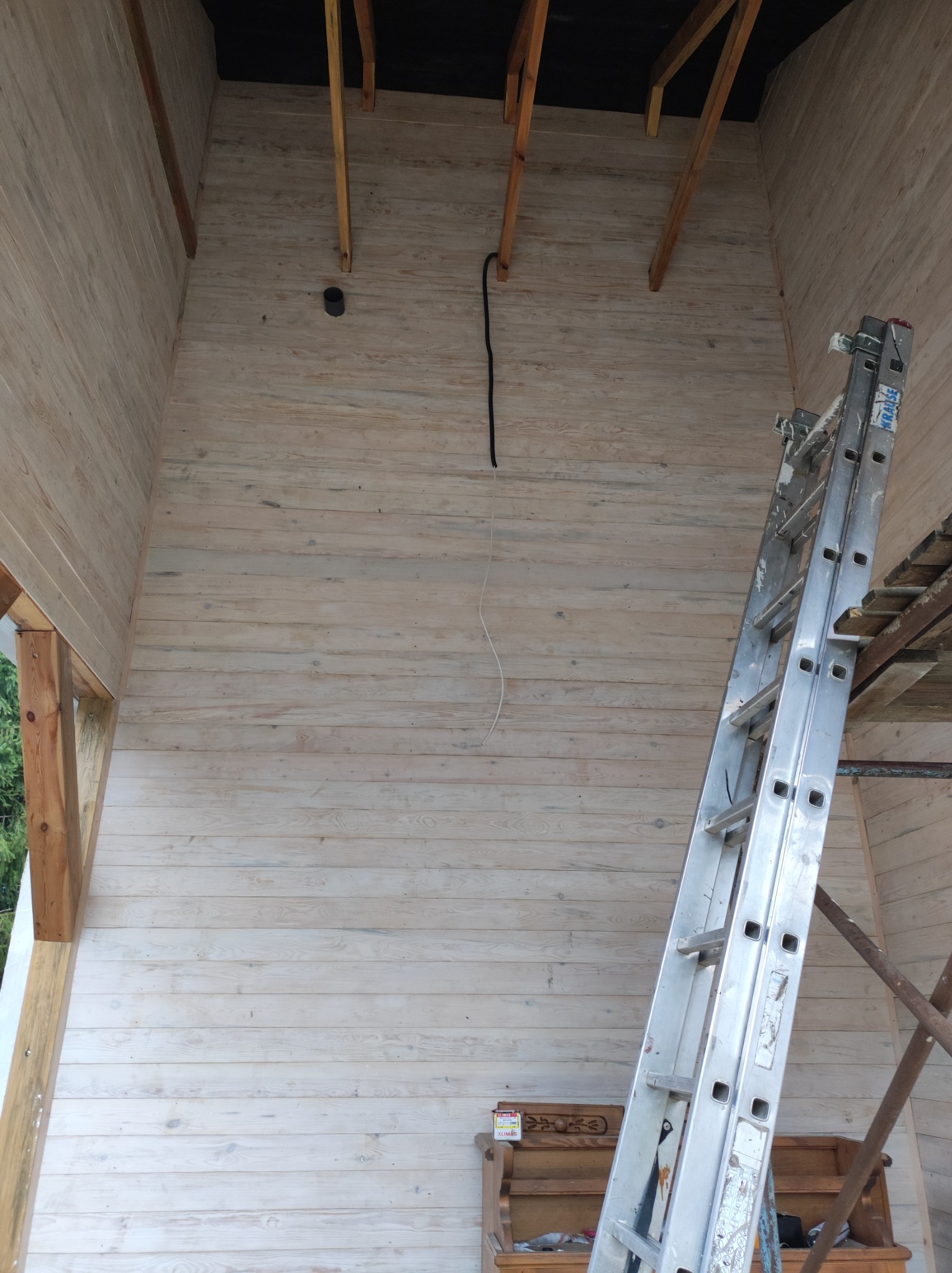 Wnętrze domu w trakcie budowy, widoczne drewniane ściany pokryte białą bejcą, drabina aluminiowa oparta o ścianę, widoczne elementy konstrukcji dachu i instalacji elektrycznej.