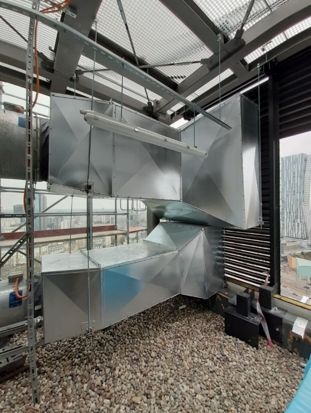 Kompleksowy system wentylacyjny z metalowymi kanałami na dachu budynku, widok na miasto w tle.