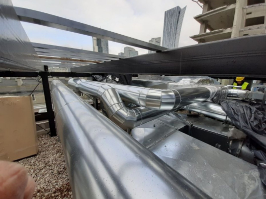 Szczegółowy widok instalacji wentylacyjnej z rurami o srebrzystej, metalicznej powierzchni, zamontowanymi na dachu budynku w trakcie budowy, z widocznymi wieżowcami w tle.