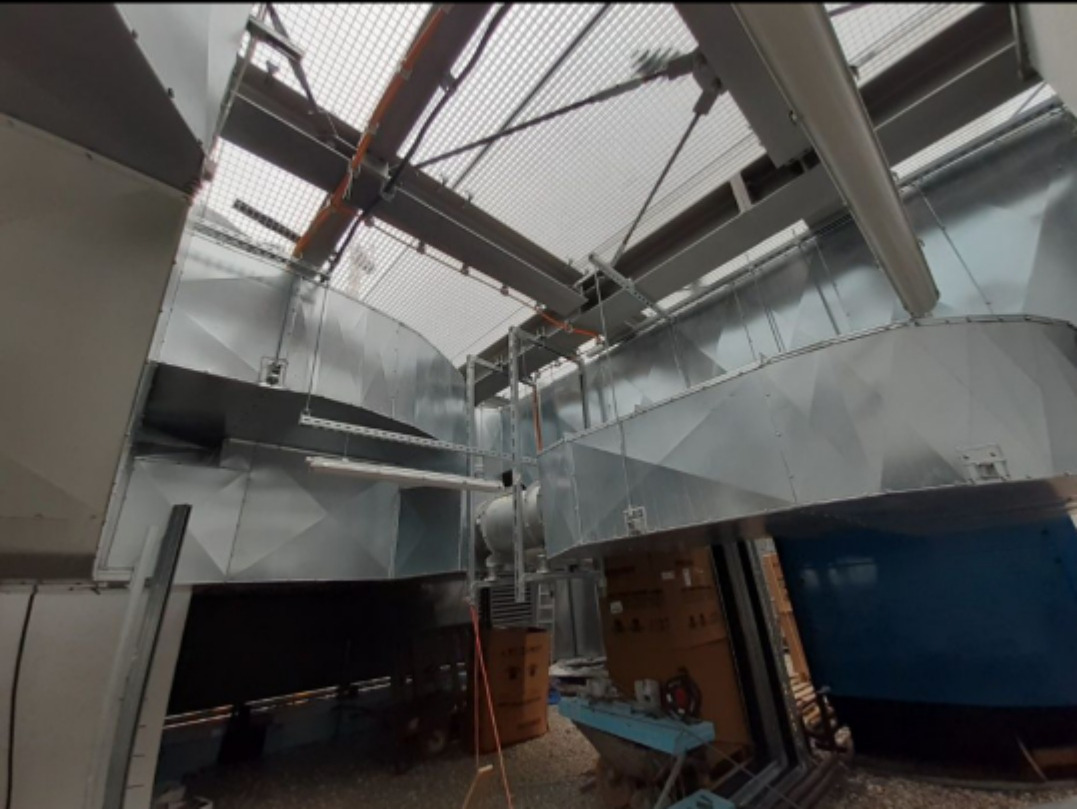 Metalowe kanały wentylacyjne pod siatkowym sufitem, widoczne elementy konstrukcyjne i instalacyjne, surowy industrialny wygląd.