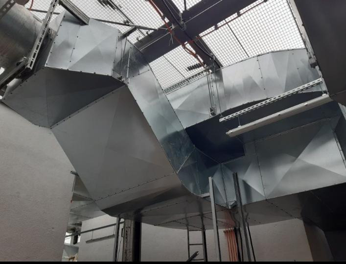 System wentylacyjny z blachy ocynkowanej, widoczny z dołu, z elementami mocującymi do sufitu i kratką wentylacyjną w tle.