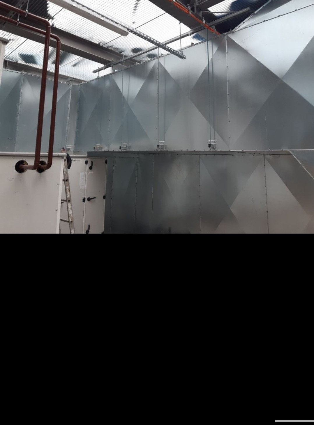 Instalacja przemysłowa z dużymi, metalowymi kanałami wentylacyjnymi o geometrycznym wzorze, widoczne rury i drabina w tle, oświetlenie sufitowe.